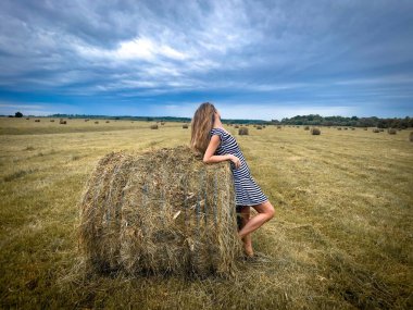 Bir tarlada samanlığın yanında duran kadın ve gökyüzünde kara fırtına bulutları.