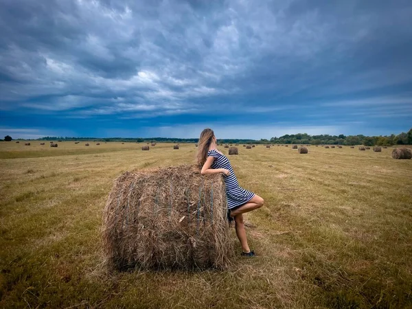 Woman standing near haystack on a field and dark storm clouds in the sky