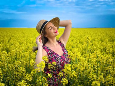 Güneşli bir günde mavi gökyüzünde kanola çiçekleri arasında hasır şapka takan bir kadın.