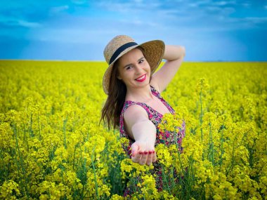 Güneşli bir günde mavi gökyüzünde kanola çiçekleri arasında hasır şapka takan bir kadın.