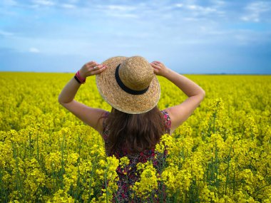 Güneşli bir günde mavi gökyüzünde kanola çiçekleri arasında hasır şapka takan bir kadın.