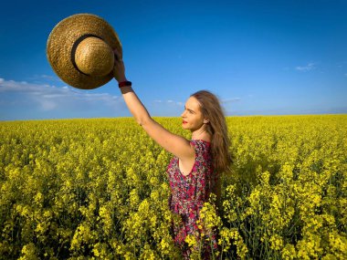 Yazlık elbiseli kadın kollarını kaldırıp kanola çiçeklerinden bir tarlada dikiliyor.