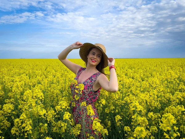 Güneşli bir günde mavi gökyüzünde kanola çiçekleri arasında hasır şapka takan bir kadın.