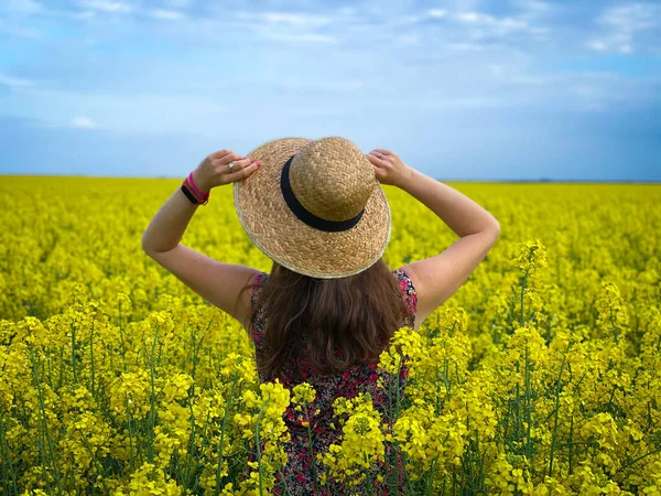 Güneşli bir günde mavi gökyüzünde kanola çiçekleri arasında hasır şapka takan bir kadın.