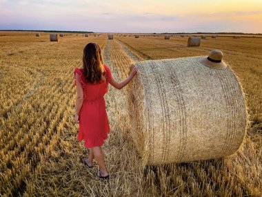 Woman in red dress with a straw hat standing near bales of hay at sunset