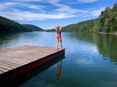 Woman in red bikini standing on a wooden pontoon near the lake in summer