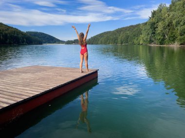 Kırmızı bikinili bir kadın yazın göl kenarında tahta bir dubanın üzerinde duruyor.