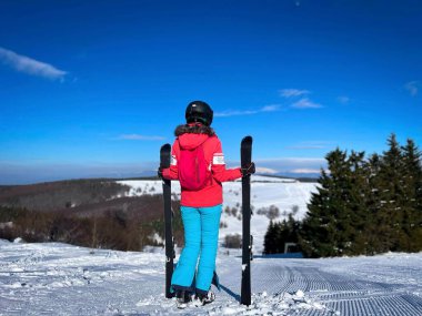 Rear view of woman wearing ski helmet and costume standing on the slope in the mountains holding her skis on a sunny winter day with clear blue sky