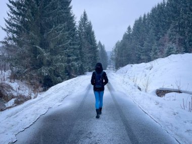 Rear view of woman with backpack and black hooded coat walking down an empty road covered in snow and surrounded by forest of coniferous trees