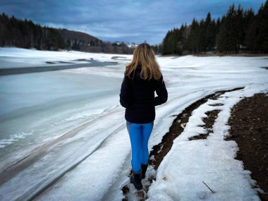Rear view of woman wearing winter clothes standing near a frozen lake surrounded by forest of coniferous trees
