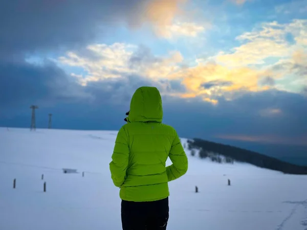 Woman in green winter coat with hood walking down a snowy road in the mountains at sunset