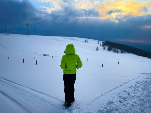 Woman in green winter coat with hood walking down a snowy road in the mountains at sunset