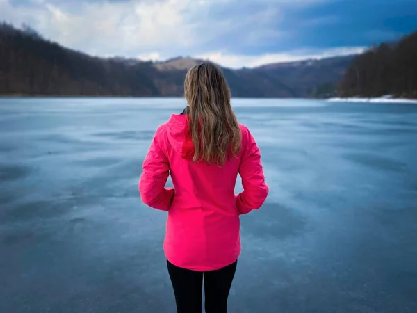 Rear view of woman with pink jacket standing near a frozen lake in the mountains