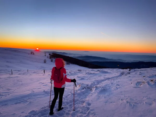 Woman with hood and trekking poles hiking alone in the mountains at sunset