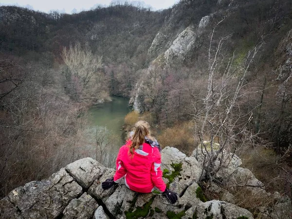 Woman sitting down on top of a cliff and looking down into the valley at a flowing river