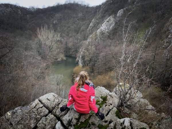 Woman sitting down on top of a cliff and looking down into the valley at a flowing river