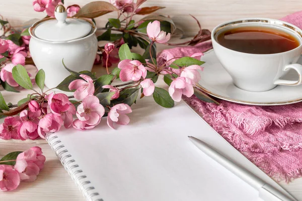 A branch of a blooming apple tree, an empty notebook for notes and a pen and a cup of tea on a wooden table. Planning the day or shopping for morning tea concept. Copy Space