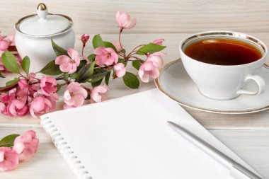 Çiçek açan elma ağacının bir dalı, notlar için boş bir defter ve ahşap bir masada bir kalem ve bir fincan çay. Günü planlamak ya da sabah çayı konsepti için alışveriş yapmak. Boşluğu Kopyala