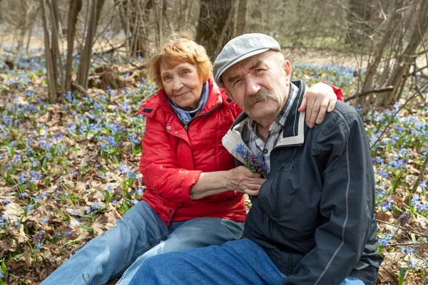 Elderly spouses relax sitting on ground in spring forest or park among blooming snowdrops — Zdjęcie stockowe