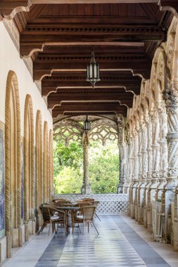 Gallery with ornate columns, wooden ceiling and marble floor in front of the entrance to the Bussaco Palace, Portugal. Tables with chairs in front of the entrance to the hotel for tourists to relax