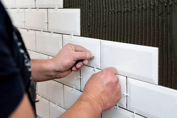 Workers hands install tile spacer between ceramic tiles. Stage of facing kitchen wall with white ceramic tiles. Repair work, construction details