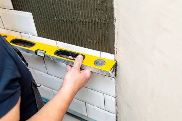 Worker hands check the evenness of the wall with a special tool for measuring the level. The stage of facing the kitchen wall with white ceramic tiles. Repair, restoration or construction work