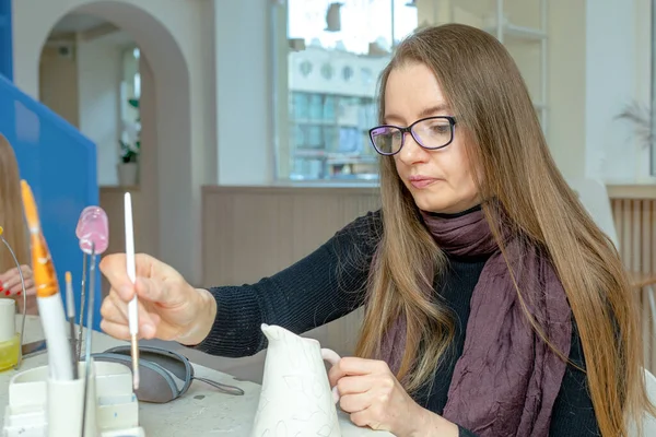 Pretty cucasian lady wearing glasses paints at a clay milk jug in a ceramic workshop. Learning new skills, improving skills, hobbies. — Stock Photo, Image