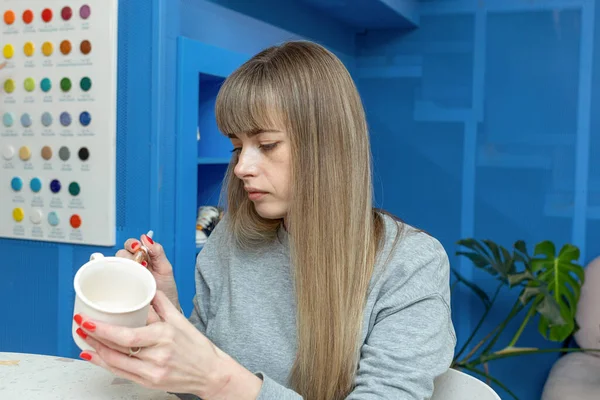 A pretty Caucasian lady paints a clay mug in a ceramic workshop. A woman holds a brush and a cup in her hands. Selective focus. — Stock Photo, Image