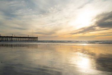Sahilde altın gün batımı. Tarihi ahşap bir Pismo Sahili İskelesi, geniş kumlu bir sahil, okyanus manzarası ve arka planda bulutlu güzel bir gökyüzü ile ufukta batan güneş.