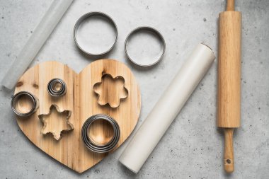 Cutters for cookie dough, parchment paper close up on wooden heart-shaped board on grey stone background, flat lay clipart