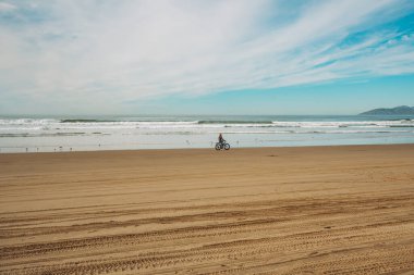 Wide sandy beach, and silhouette of a cyclist. Pacific ocean and cloudy sky on the background.