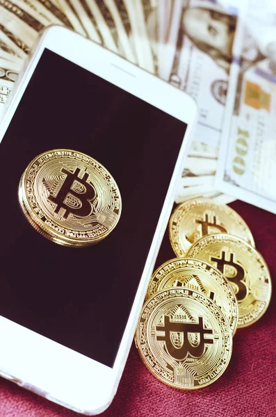 Oro físico Bitcoin Moneda contra billetes de dólar y teléfono inteligente sobre un fondo morado . — Foto de Stock