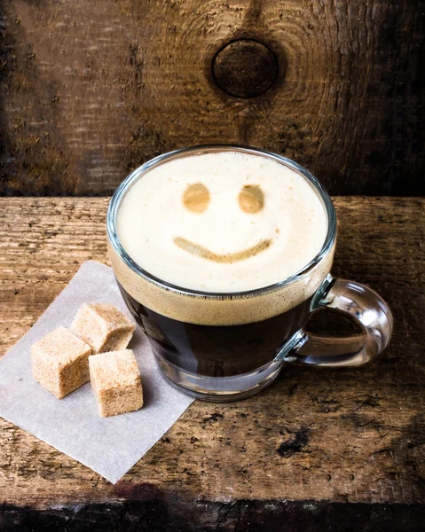 Small cup of espresso coffee in glass with smile pattern on old wooden table. — Stock Photo, Image