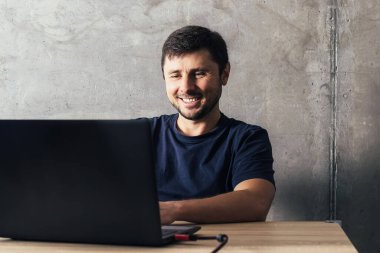 Ofis masasında oturan yakışıklı, sakallı adam, modern ofisin beton duvarına karşı dizüstü bilgisayarla çalışıyor.