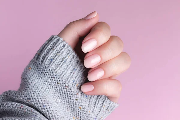 Gri örgü süveterli, doğal, güzel manikürlü, pembe arka planda pembe tırnaklı bir kadın eli. Çivi bakımı kavramı