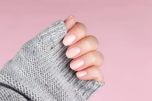 Female hand in gray knitted sweater with beautiful natural manicure - pink nude nails on pink background. Nail care concept