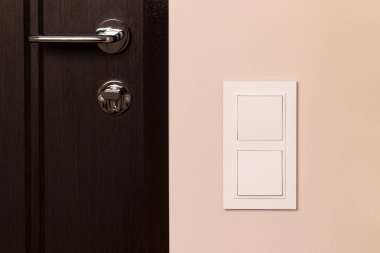 Modern apartmandaki ahşap kapının yanındaki bej duvarda beyaz elektrik düğmesi var.