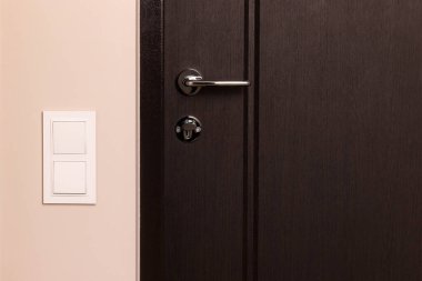 Modern apartmandaki ahşap kapının yanındaki bej duvarda beyaz elektrik düğmesi var.