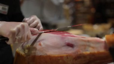 Siyah önlüklü ve beyaz eldivenli bir şef bir restoranda domuz budu kesiyor. Şef bir dilim jamon kesiyor.