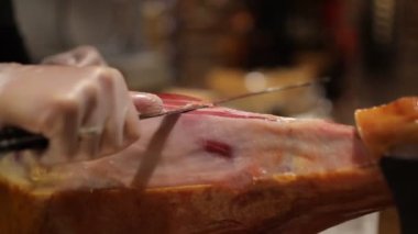 Siyah önlüklü ve beyaz eldivenli bir şef bir restoranda domuz budu kesiyor. Şef bir dilim jamon kesiyor.