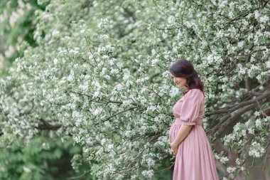 Doğada çiçek açan bir ağacın yanında duran güzel, hamile bir kadın..
