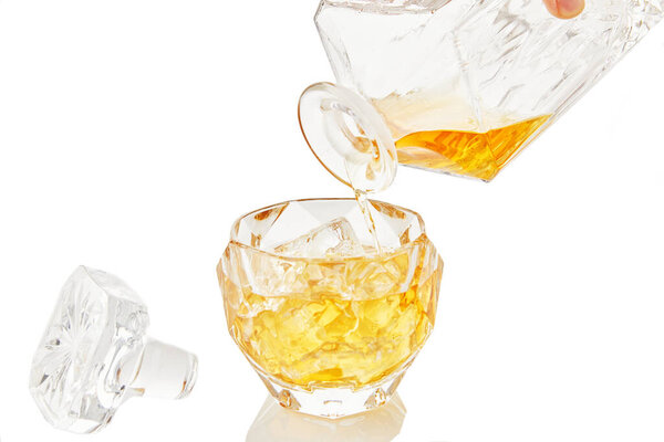 Whiskey from decanter is poured into glass of whiskey with ice on white background with reflection.