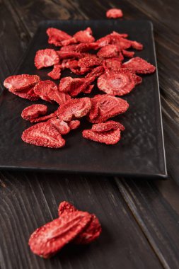 Çilek cipsleri siyah seramik tepside, koyu ahşap arka planda. Yüzde yüz doğal, dondurulmuş çilek. Sağlıklı sebze cipsi