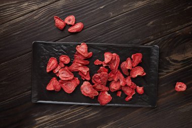 Çilek cipsleri siyah seramik tepside, koyu ahşap arka planda. Yüzde yüz doğal, dondurulmuş çilek. Düz yatıyordu. Sağlıklı sebze cipsi