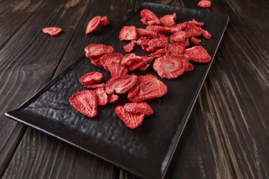 Çilek cipsleri siyah seramik tepside, koyu ahşap arka planda. Yüzde yüz doğal, dondurulmuş çilek. Sağlıklı sebze cipsi
