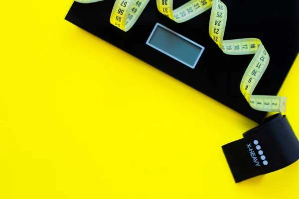 Una bilancia di peso, un nastro nero per il fitness e un nastro di misurazione giallo si trovano su uno sfondo giallo sul lato della foto. vista dall'alto, disposizione piatta, spazio copia, isolare. — Foto Stock