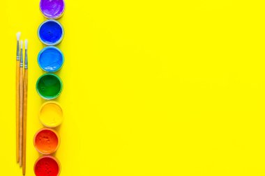 Boya kutuları yan çizgide duruyor, renkler gökkuşağının sırasına göre sergileniyor, yakınlarda sarı bir zemin üzerinde boya fırçaları var. Üst görünüm, düz görünüm, kopyala boşluk, izole et