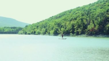 Bir kız nehirde bir tahtanın üzerinde yüzüyor, arka planda ağaçlarla kaplanmış dağlar var.. 