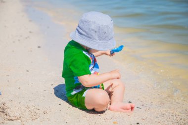 Su kenarındaki kumların üzerinde panama şapkalı küçük bir çocuk oturuyor ve kürekle oynuyor.. 