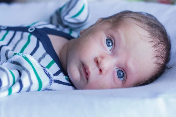 Un bebé pequeño con los ojos azules en un traje a rayas se encuentra en una almohada blanca y se ve ligeramente a un lado del marco. — Foto de Stock